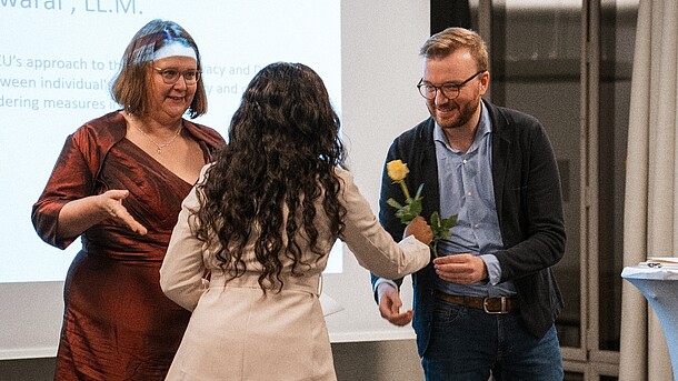 Prof. Dr. Margrit Seckelmann und Florian Mäder überreichen einer Absolventin das Zeugnis und eine gelbe Rose.